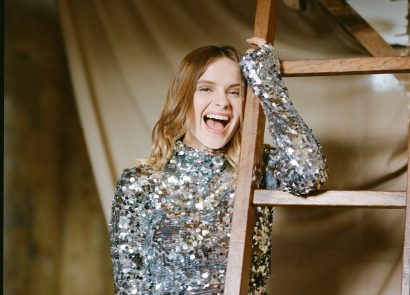Gabrielle Aplin smiles in a silver sequin dress holding onto a wooden ladder