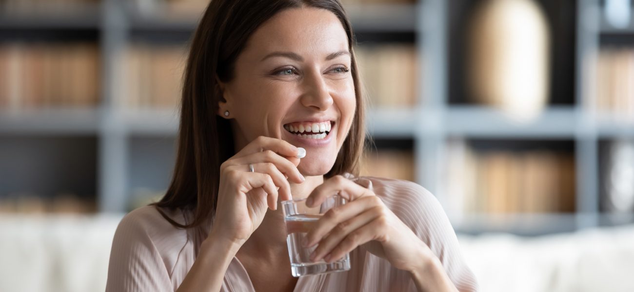 Woman smiling, holding pill and glass of water, after beating bloating