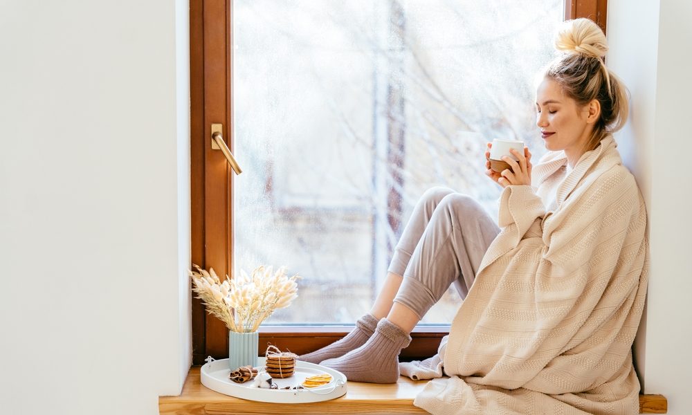 Happy blond woman with bunch hairstyle warming and cover knitted plaid enjoying in her coffee time by the window in cold winter day