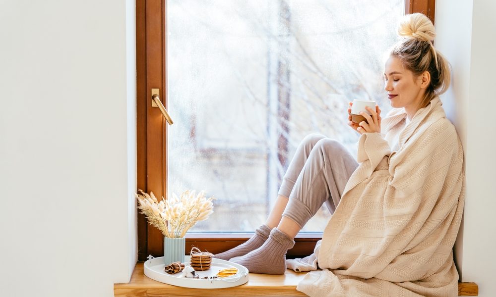 Happy blond woman with bunch hairstyle warming and cover knitted plaid enjoying in her coffee time by the window in cold winter day