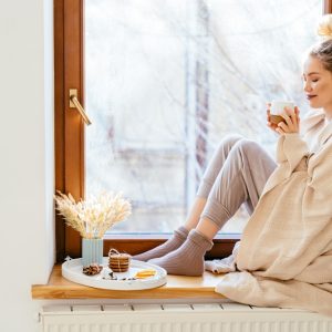 Happy blond woman with bunch hairstyle warming and cover knitted plaid enjoying in her coffee time by the window in cold winter day