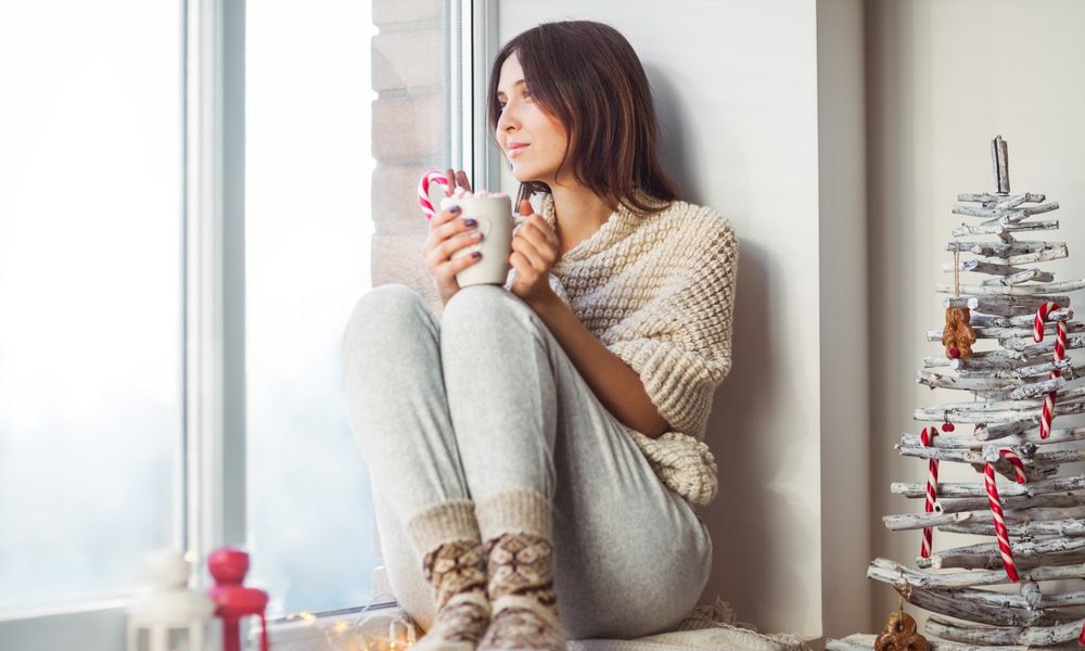 Happy beautiful woman drinking hot coffee sitting on window sill in christmas decorated home