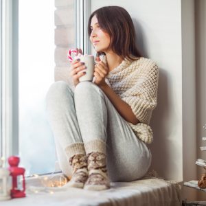 Happy beautiful woman drinking hot coffee sitting on window sill in christmas decorated home