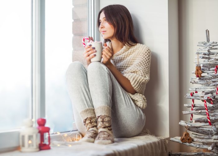 Happy beautiful woman drinking hot coffee sitting on window sill in christmas decorated home