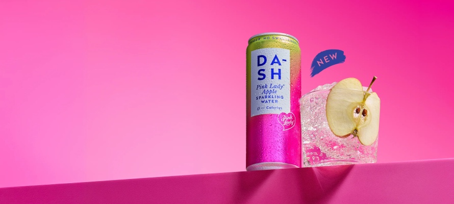 A can of DASH Pink Lady Apple Sparkling Water next to a glass with ice and a slice of apple on a pink background