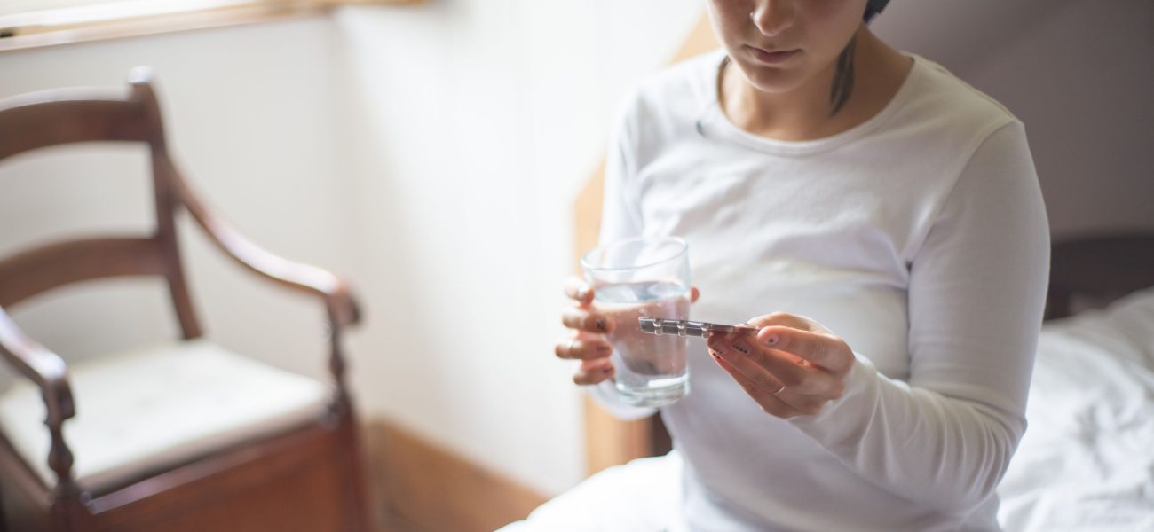 A woman sitting on a bed holding a glass of water while looking a pack of medicinal tablets