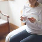 A woman sitting on a bed holding a glass of water while looking a pack of medicinal tablets