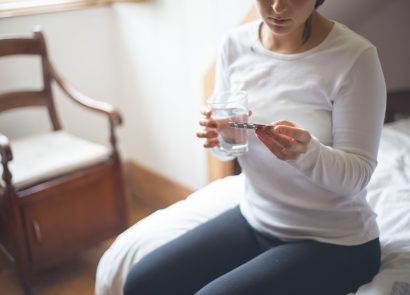 A woman sitting on a bed holding a glass of water while looking a pack of medicinal tablets