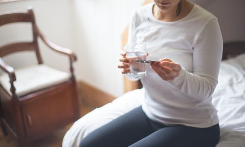 A woman sitting on a bed holding a glass of water while looking a pack of medicinal tablets