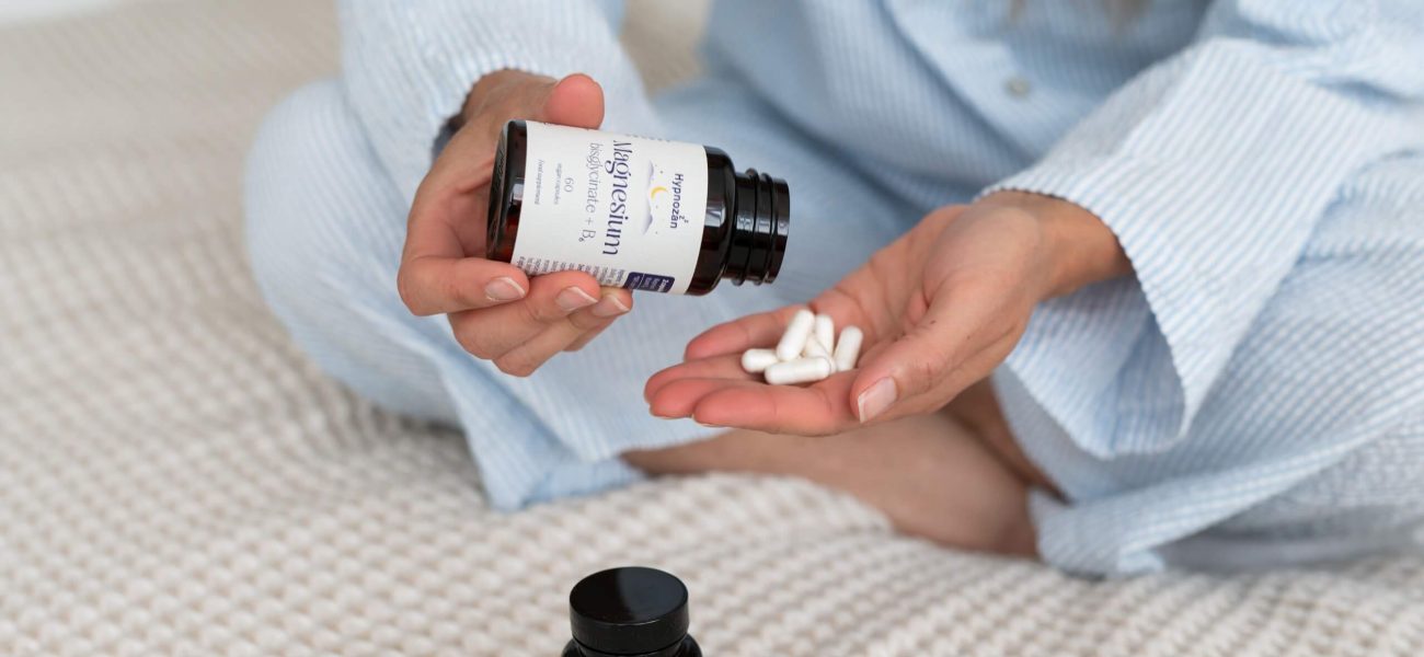 A woman tipping Hypnozan capsules into her hand as she sits on a bed; another jar of Hynozan is beside her on the duvet
