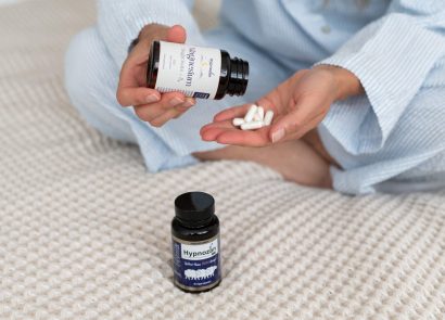A woman tipping Hypnozan capsules into her hand as she sits on a bed; another jar of Hynozan is beside her on the duvet