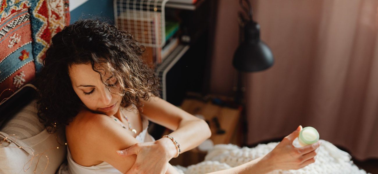 A dark-haired woman sitting on her bed apply CBD cream to her arm