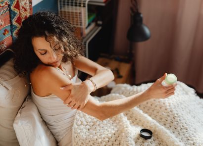A dark-haired woman sitting on her bed apply CBD cream to her arm