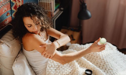 A dark-haired woman sitting on her bed apply CBD cream to her arm