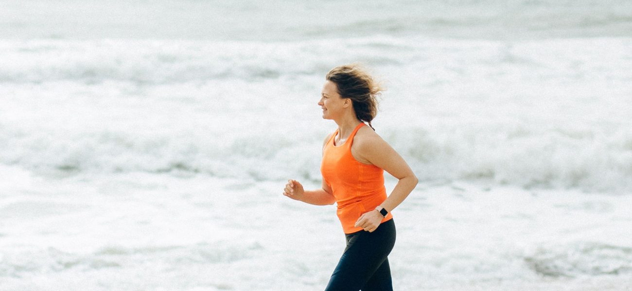 A middle-aged woman jogging on a beach with the sea behind her