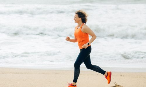 A middle-aged woman jogging on a beach with the sea behind her