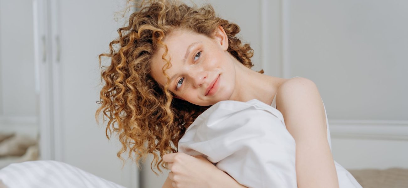 A smiling woman sitting on a bed with a bedsheet wrapped around her