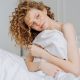 A smiling woman sitting on a bed with a bedsheet wrapped around her