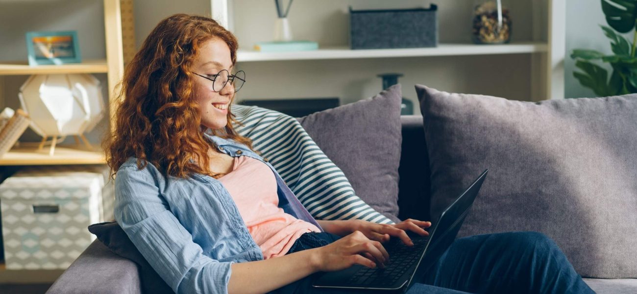 A woman with red hair wearing glasses sitting on a sofa using a laptop computer