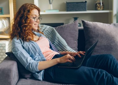 A woman with red hair wearing glasses sitting on a sofa using a laptop computer