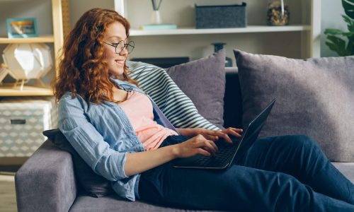 A woman with red hair wearing glasses sitting on a sofa using a laptop computer