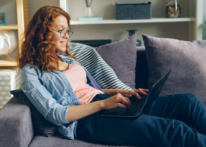 A woman with red hair wearing glasses sitting on a sofa using a laptop computer