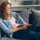 A woman with red hair wearing glasses sitting on a sofa using a laptop computer